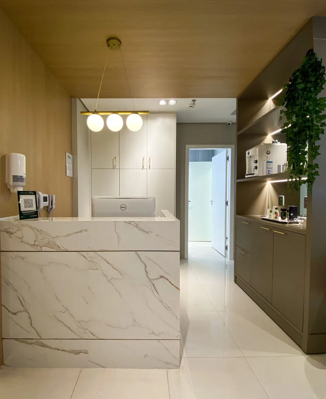 Balcão de recepção em mármore branco, com luminárias suspensas e corredor de acesso aos consultórios ao fundo.