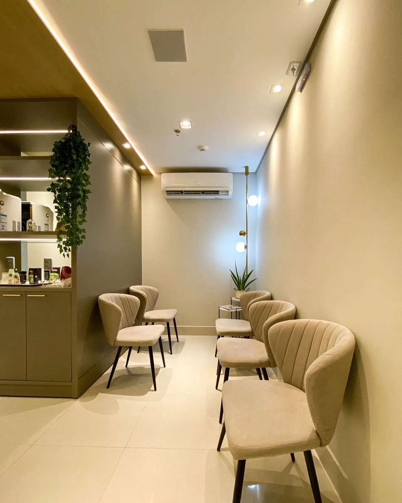 Sala de espera com poltronas estofadas claras dispostas ao longo da parede, iluminação indireta e plantas.